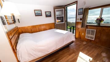 A bedroom with a wooden bed, a window with a view of the sea, and a door leading outside.