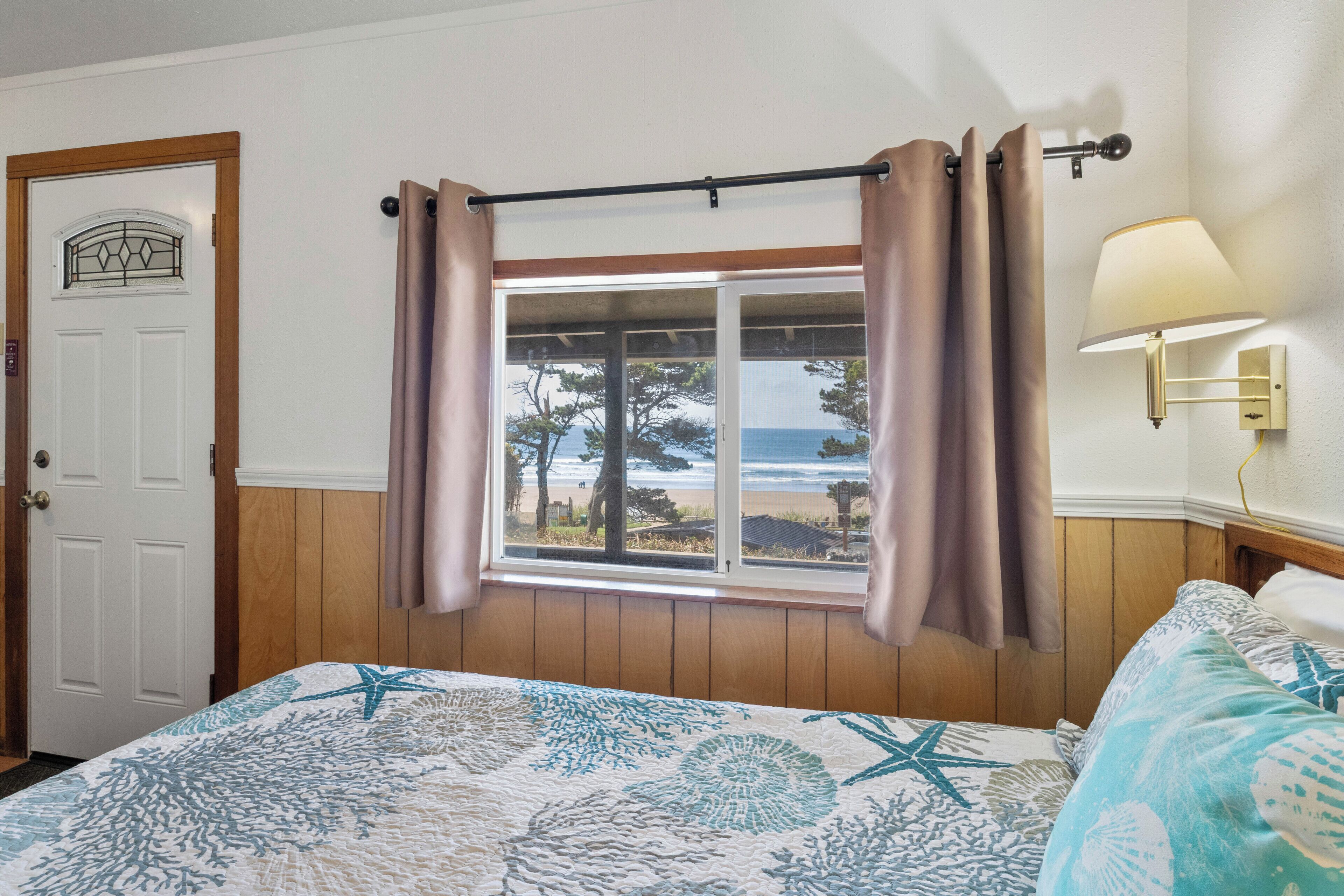 A bedroom with a bed, a window with curtains, a door, and a wall-mounted lamp.