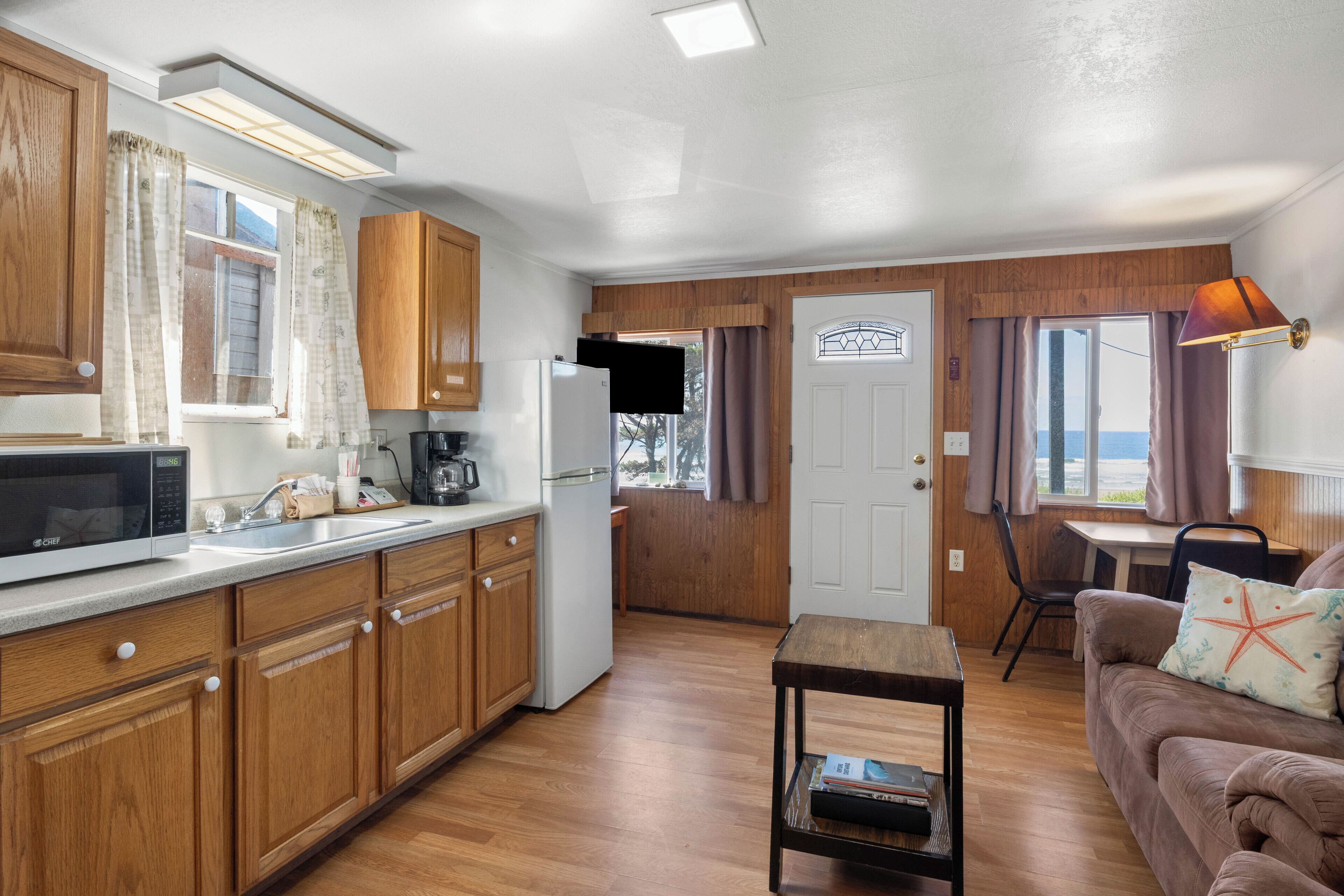Cocina compacta con armarios de madera, microondas y refrigerador. Hay una zona de comedor con una mesa y sillas, y un espacio de sala con un sofá y un cojín con forma de estrella de mar.