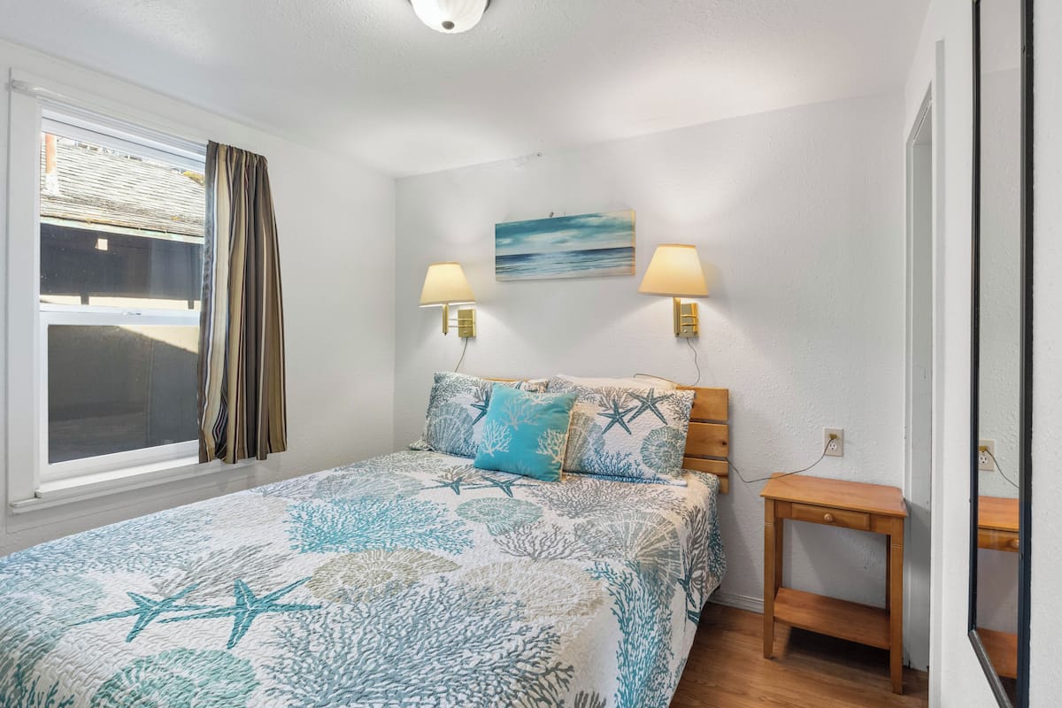 A bedroom with a bed, two wall-mounted lamps, a painting, a wooden nightstand, and a window with curtains.