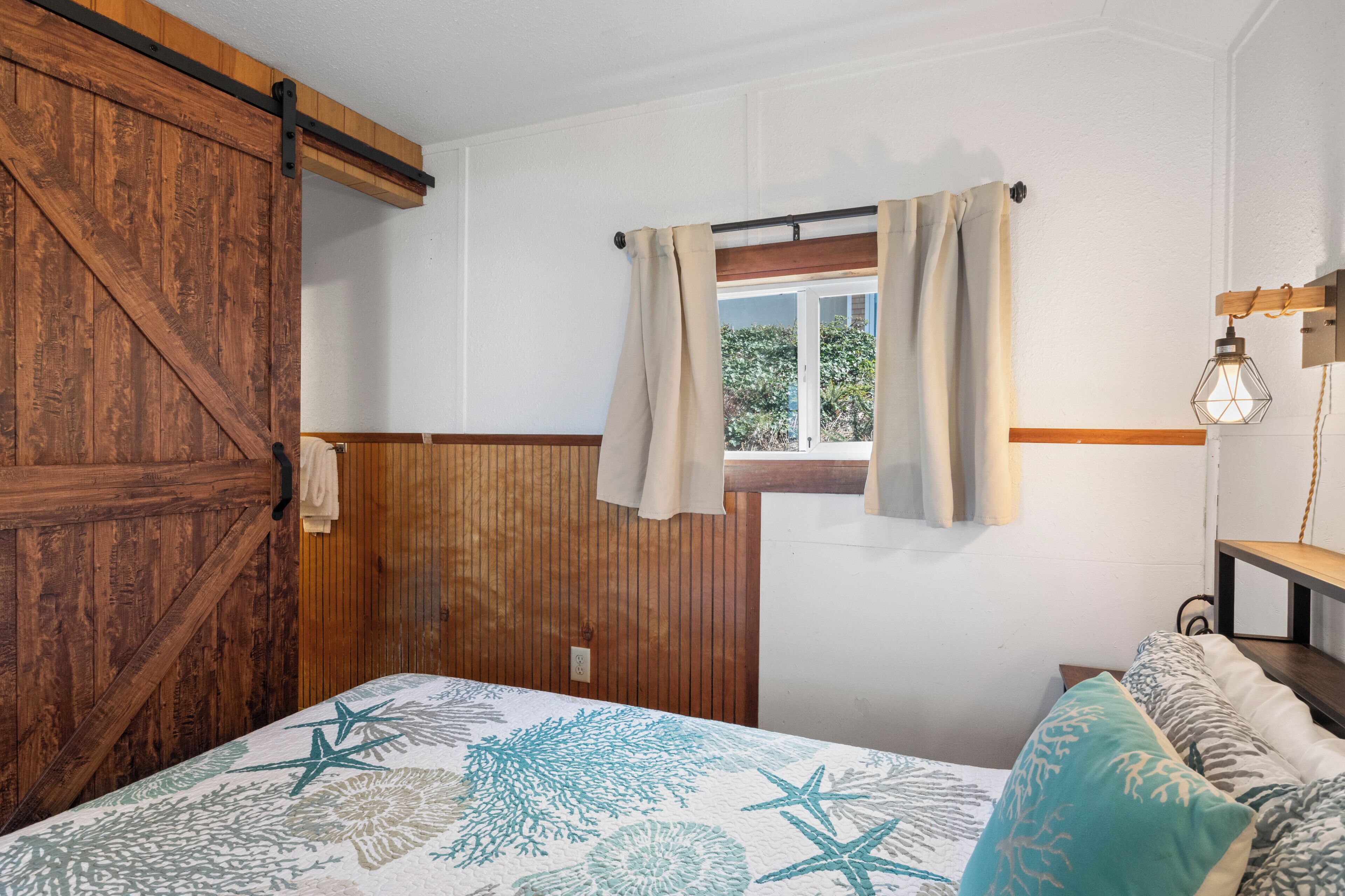 Une chambre avec une armoire en bois, un lit recouvert d’une couvre-lit à motifs, une fenêtre avec des rideaux et une lampe de chevet.