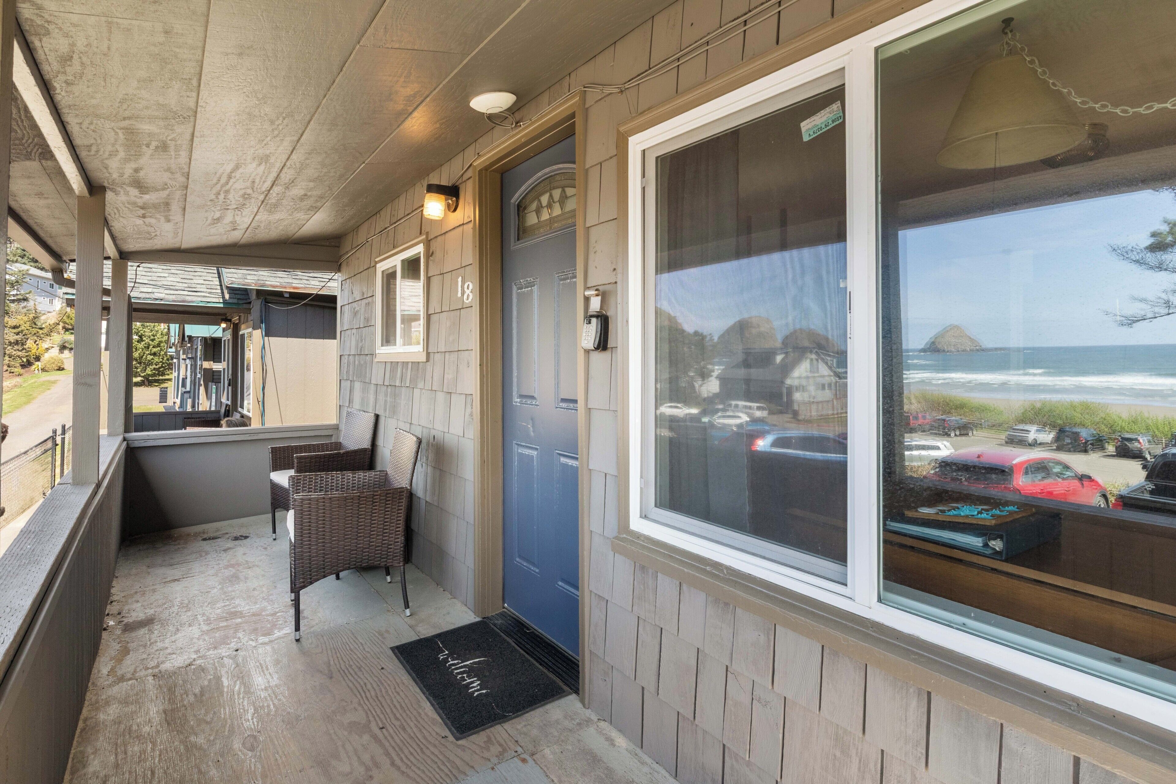 A covered porch with wicker chairs, a blue door, and a view of the ocean and parked cars.