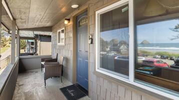 A covered porch with wicker chairs, a blue door, and a view of the ocean and parked cars.