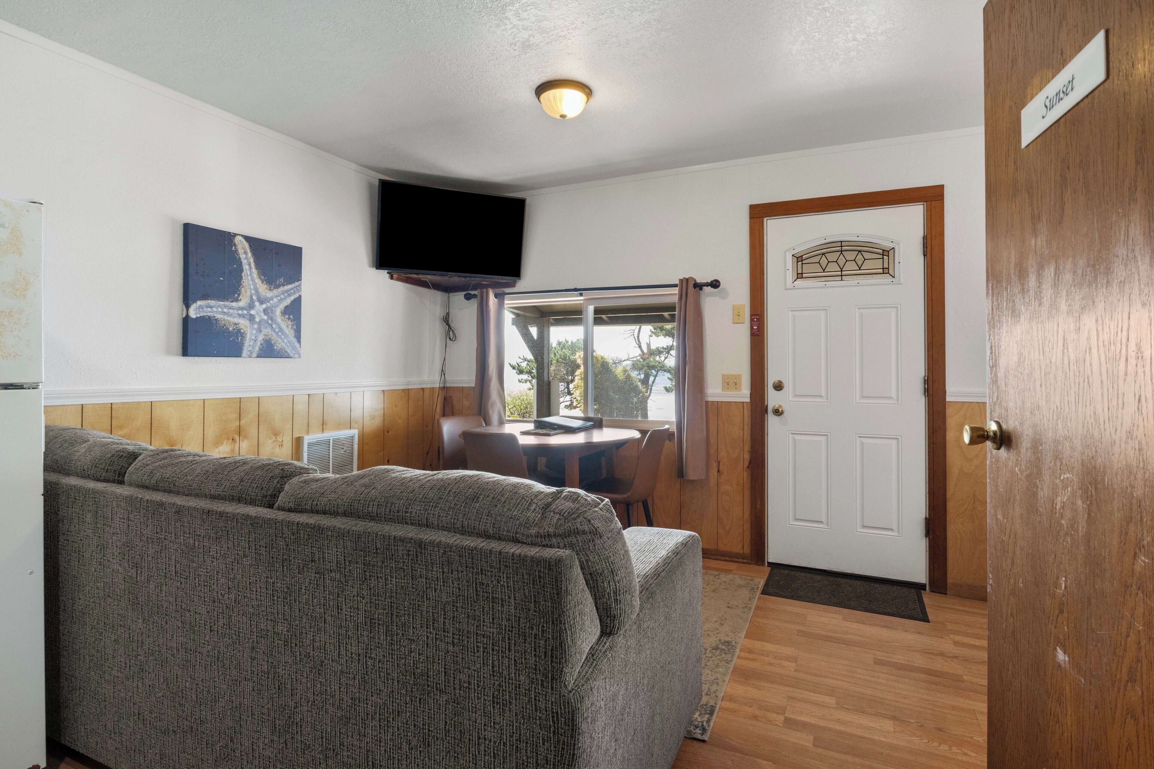 Una habitación con sofá, televisor, una puerta con un letrero y una ventana con cortinas.