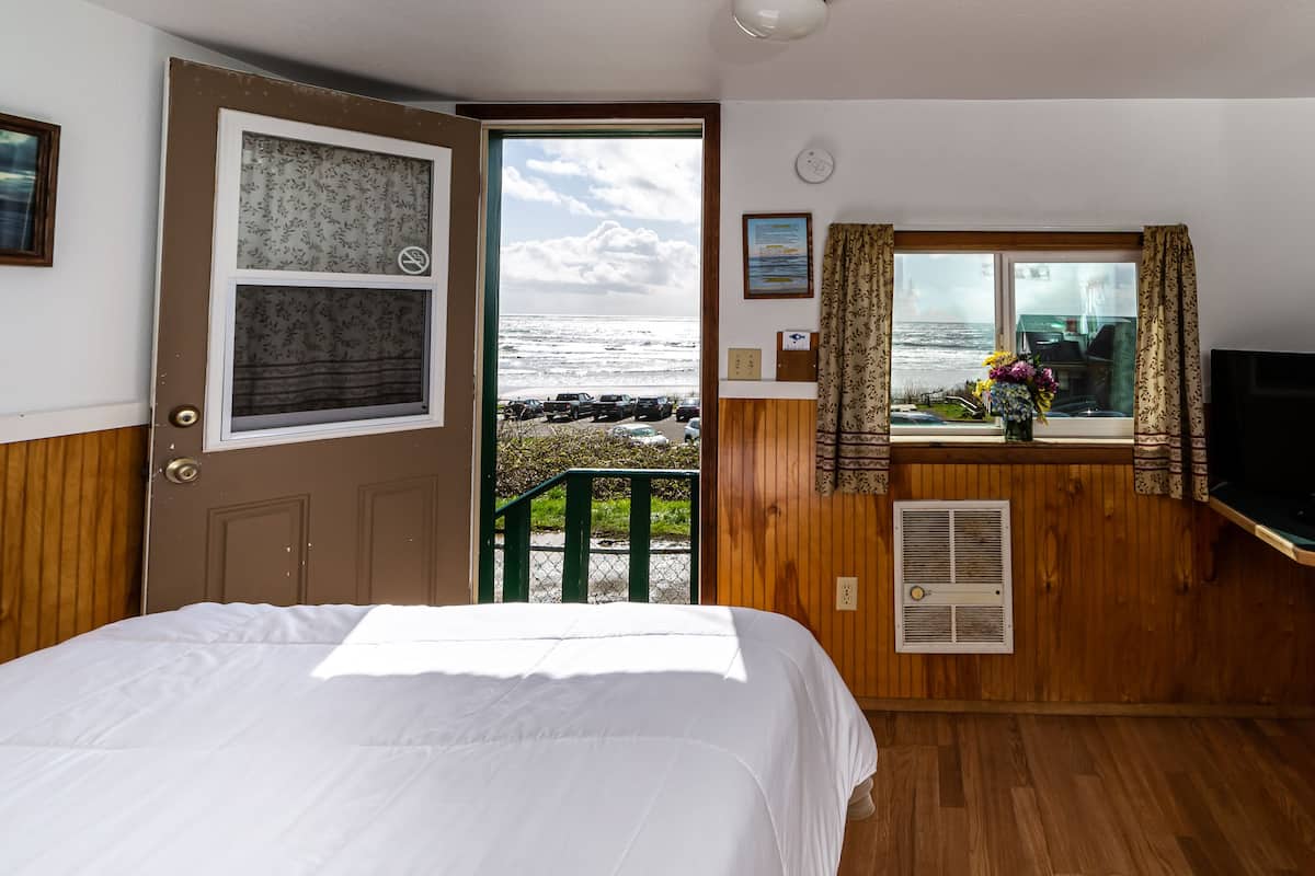 A room with a bed, a window with curtains, a small table with a vase of flowers, and a view of the ocean.