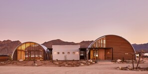 A modern building with a curved roof and large windows, surrounded by a desert landscape with mountains in the background.