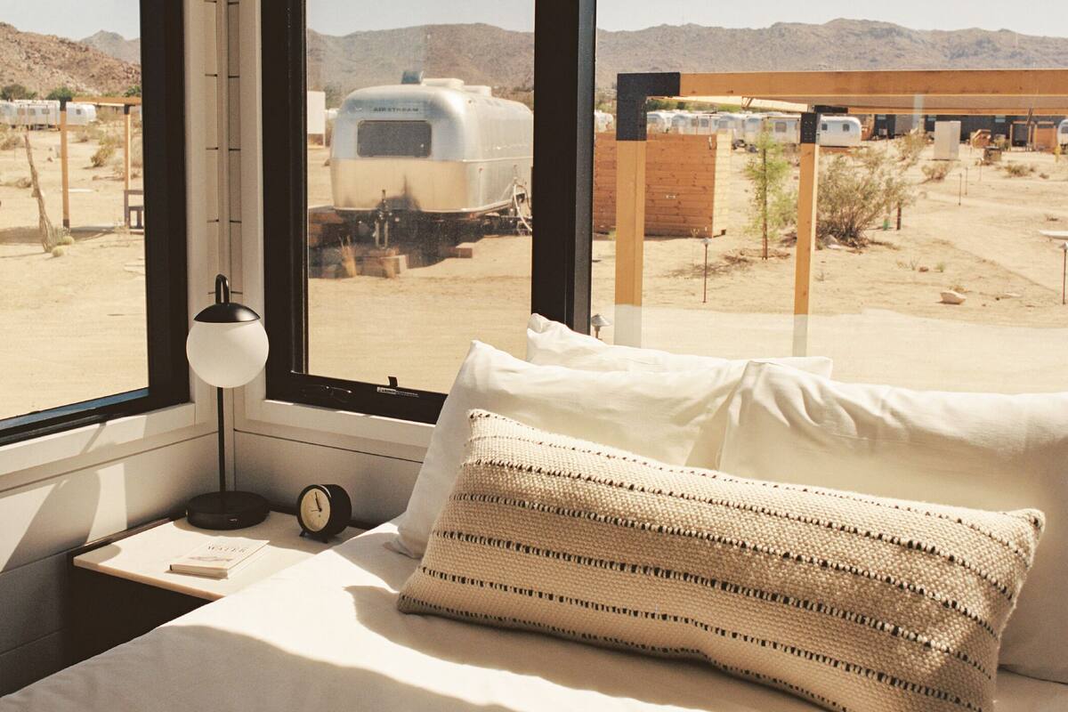 A bedroom with a bed, a bedside table with a lamp, and a view of a desert landscape with trailers.
