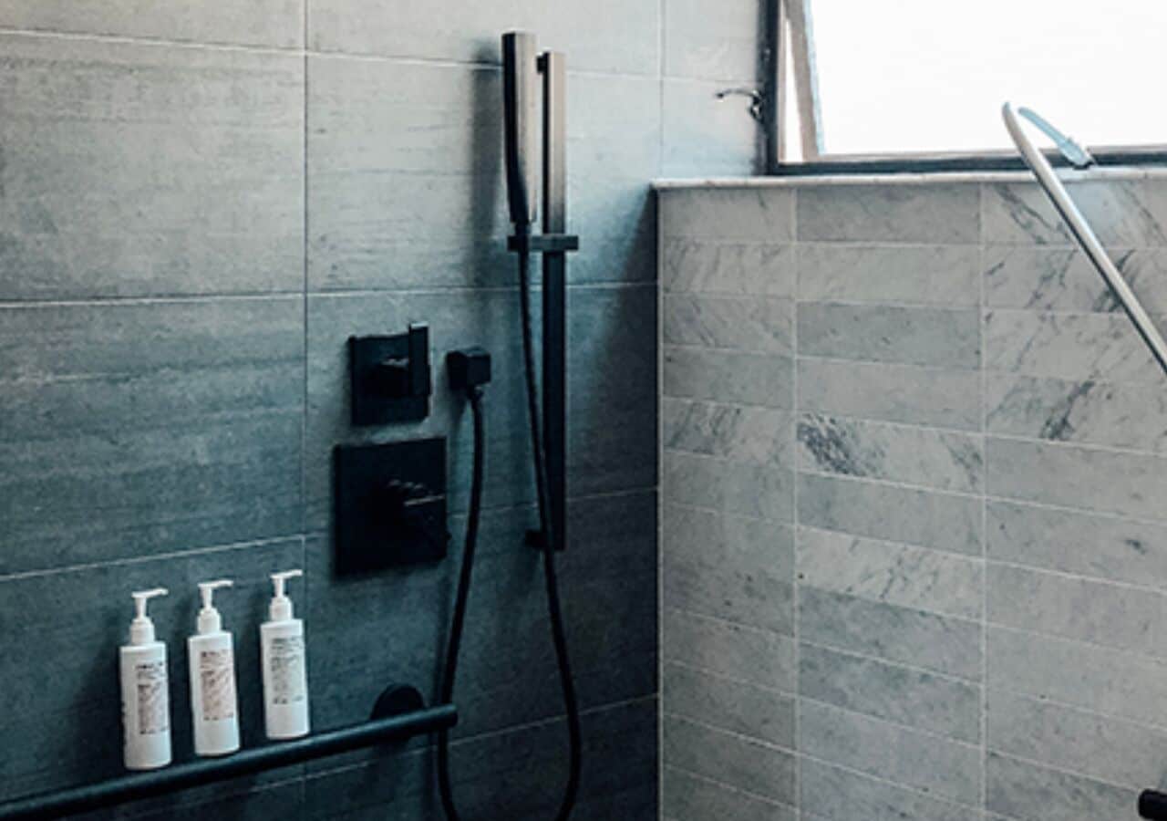 A bathroom with grey tiles, a shower, and a shelf with toiletries.