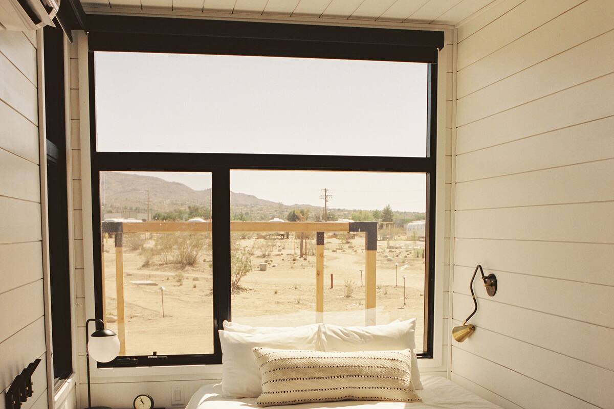 A bedroom with a large window, a bed, a bedside table with a lamp, and a view of a desert landscape.