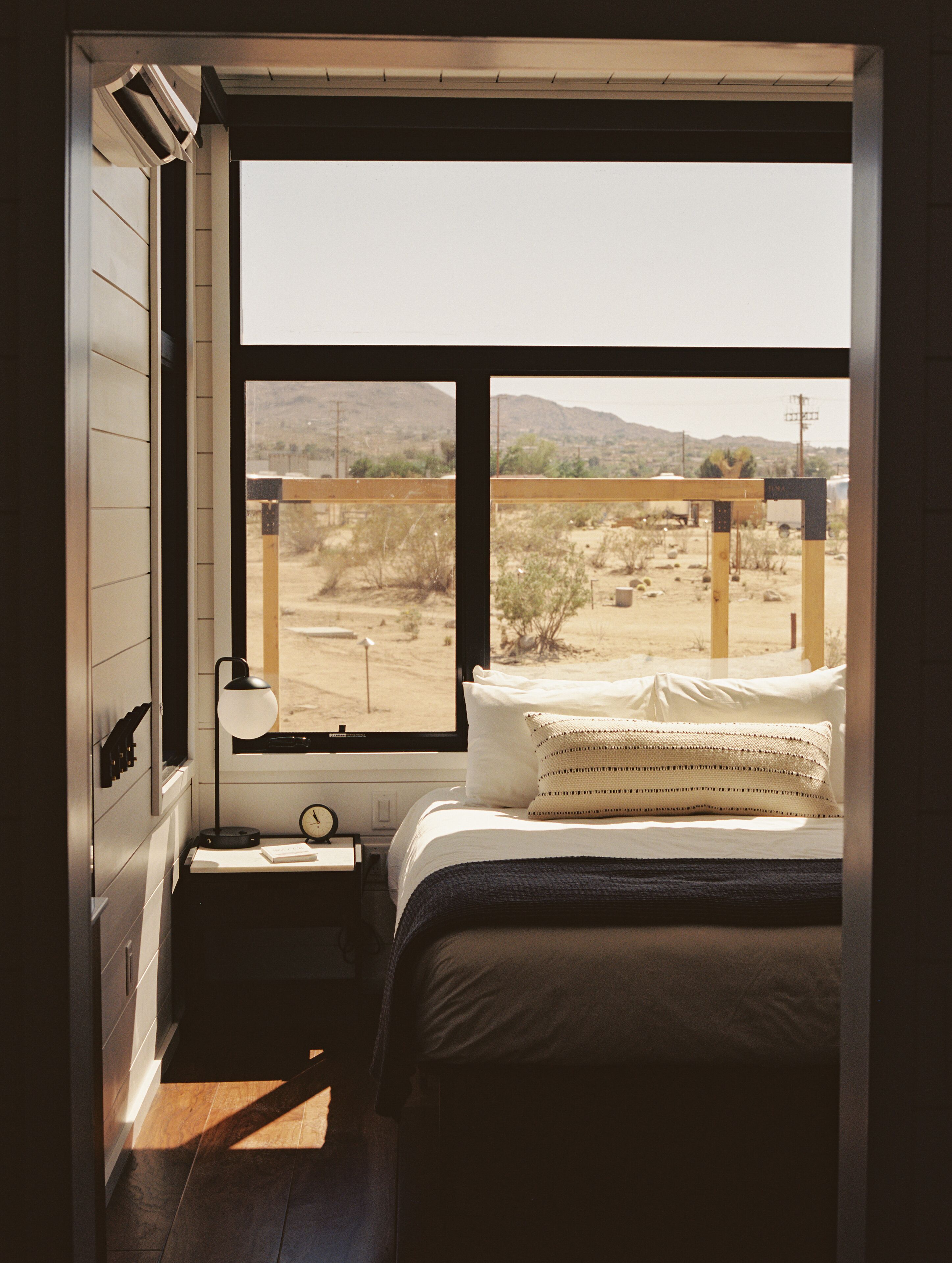 A bedroom with a large window, a bed with white bedding, a bedside table with a lamp, and a view of a desert landscape.