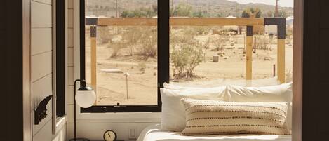 A bedroom with a large window, a bed with white bedding, a bedside table with a lamp, and a view of a desert landscape.