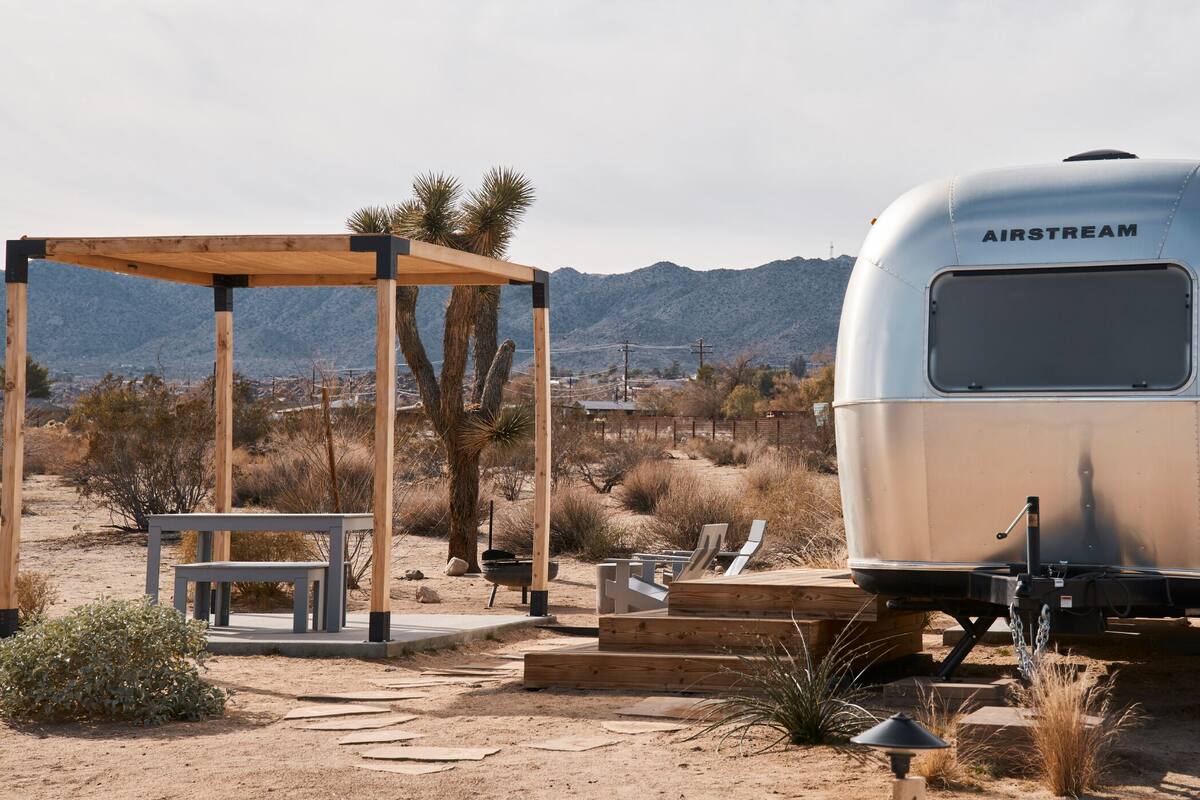 A silver AIRSTREAM trailer with a covered outdoor seating area and a view of mountains in the background.