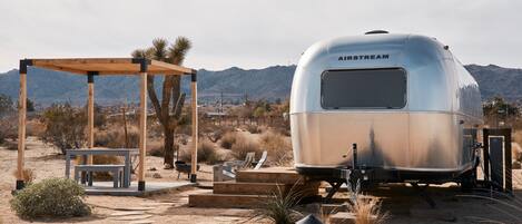 Une caravane AIRSTREAM argentée, dotée d’un espace salon extérieur abrité et offrant une vue sur les montagnes en arrière-plan.