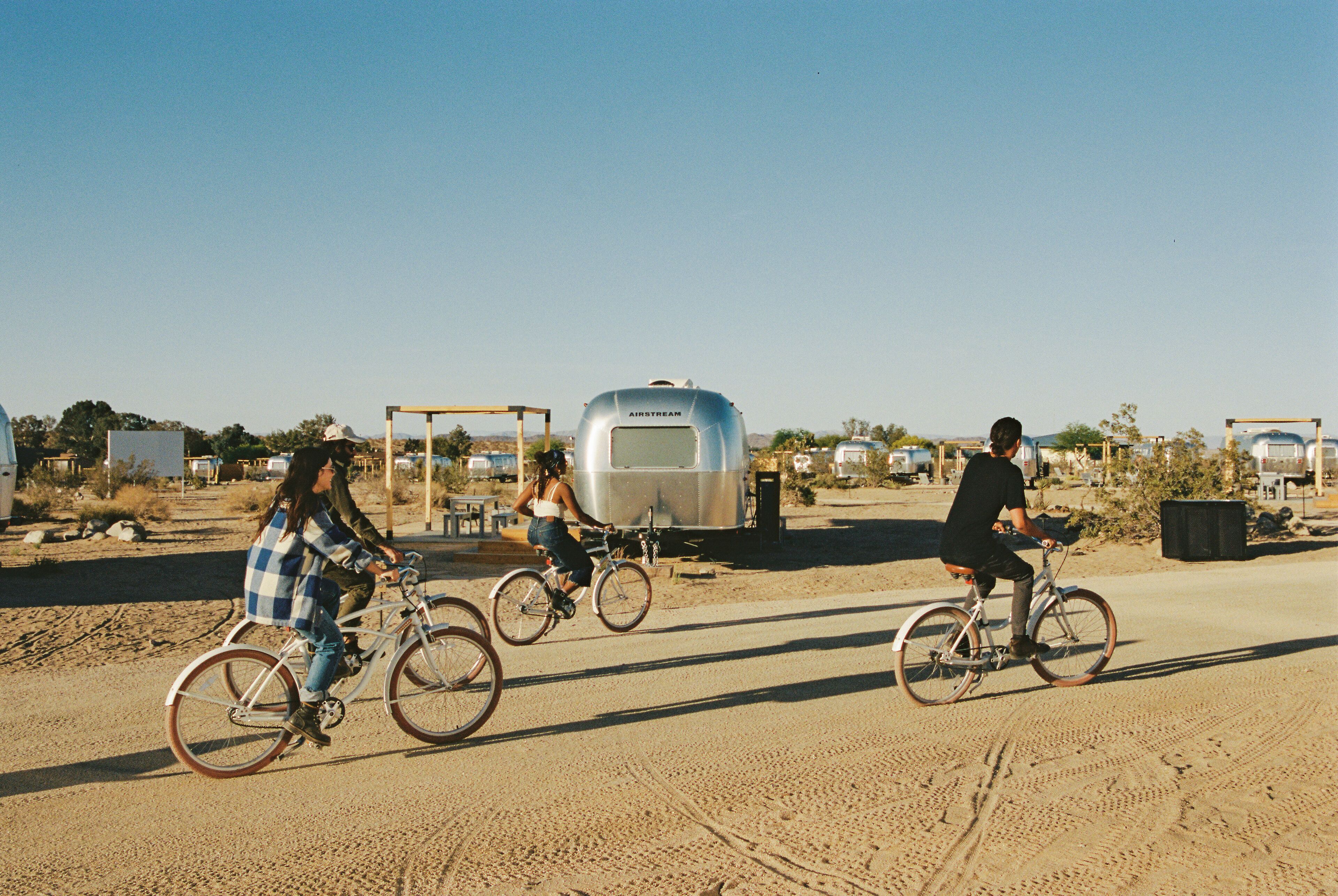 Three people riding bicycles on a dirt road with a trailer in the background.