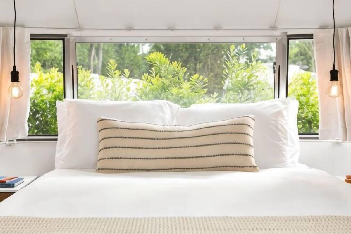A bed with white bedding and striped pillows, a window with curtains, and a view of greenery outside.