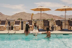 A swimming pool with lounge chairs and umbrellas, surrounded by a wooden fence and desert hills in the background.