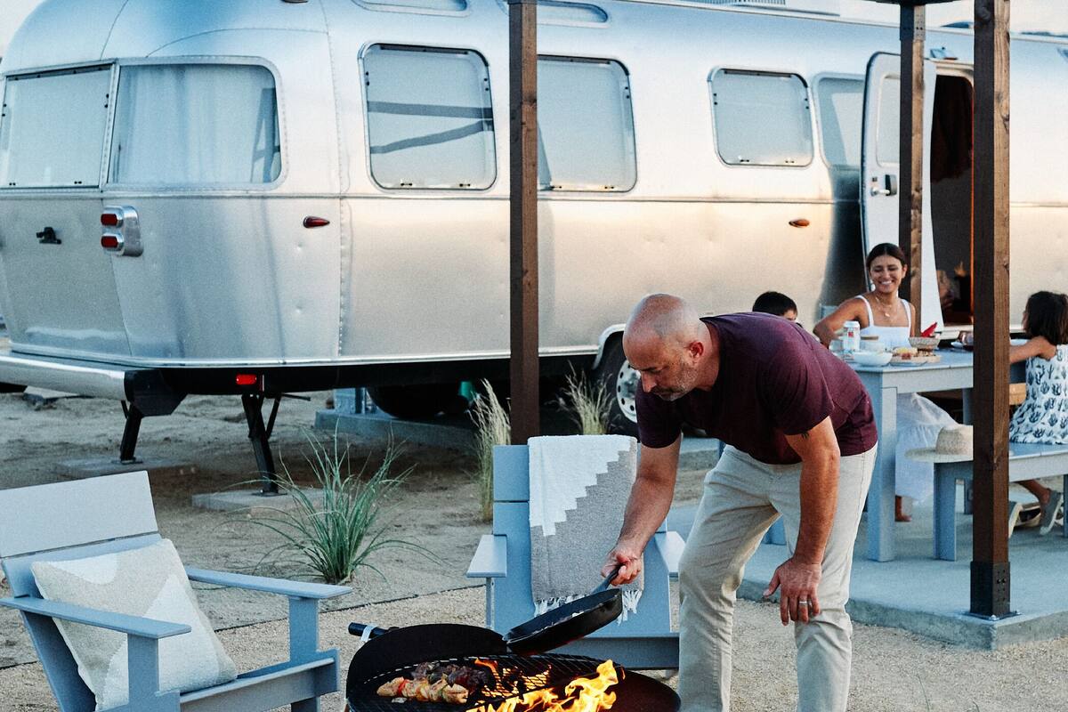 A man grilling outdoors with a trailer and a dining area in the background.