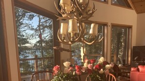 A dining area with a round wooden table, chairs, a chandelier, and a view of trees through large windows.