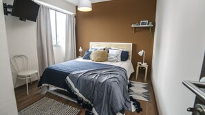 A neatly arranged bedroom with a bed, bedside tables, a TV mounted on the wall, and a window with curtains.