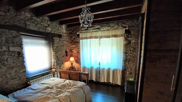 Een landelijke slaapkamer met een stenen muur, een bed met een geborduurde spreideken, een houten nachtkastje met een lamp en een kroonluchter.