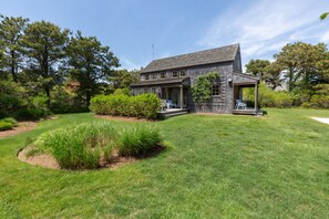 A two-story house with a covered porch, surrounded by a well-maintained lawn and various shrubs. - Pond View Cottage- Luxury home near Miacomet beach (Nantucket)
