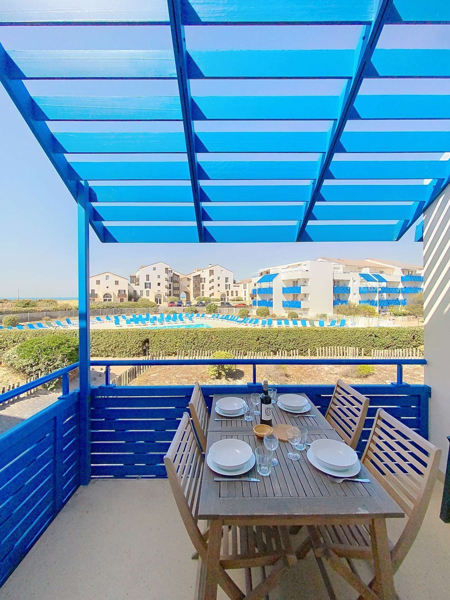 A balcony with a blue pergola, a wooden table set for dining, and a view of buildings and a pool in the background.