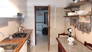 Une petite cuisine équipée d’une cuisinière à gaz, d’un évier et d’un plan de travail sur lequel se trouve une bouilloire. Une porte donne accès à une chambre à coucher avec un lit et un mur décoré de motifs.