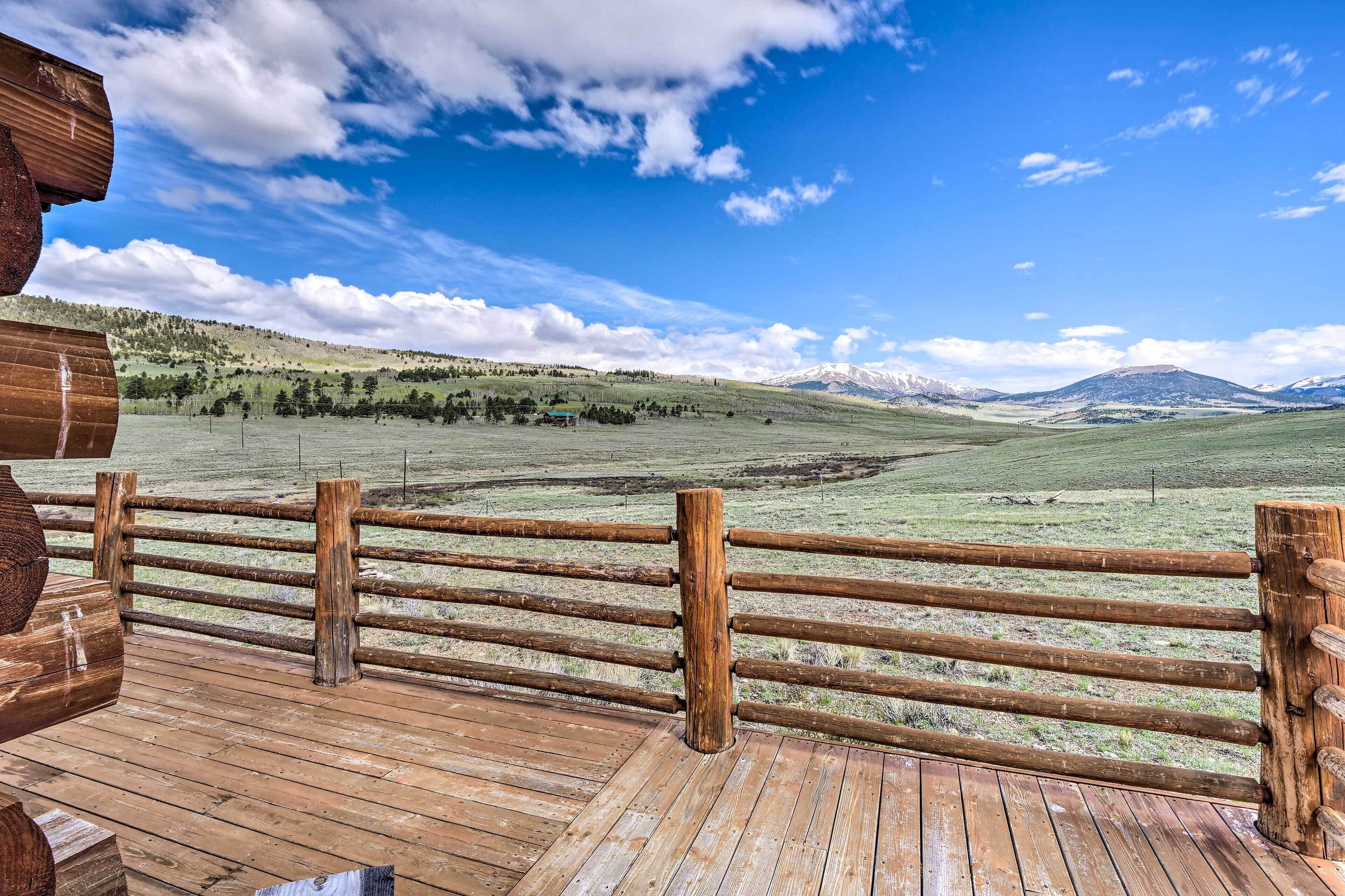 Une terrasse en bois, entourée d’une clôture également en bois, donnant sur un champ verdoyant avec des montagnes qui se dessinent à l’horizon.