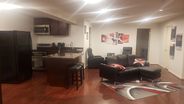 A modern kitchen with a central island, a dining area with a black leather sofa, and a living room with a black leather sofa and a red and white patterned rug.