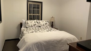 Een keurig opgemaakt bed met een witte dekbedovertrek en gestreepte kussens, een nachtkastje met een lamp, en een raam met uitzicht op de omgeving.
