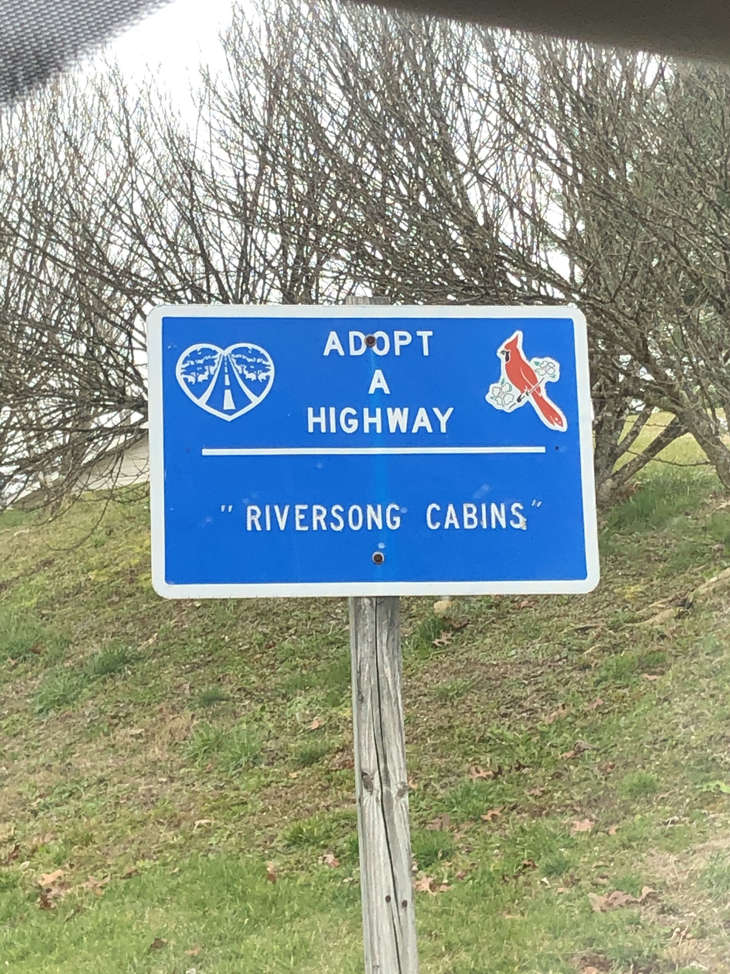 A blue sign with white text and a red bird graphic, advertising 'Riversong Cabins' as a place to adopt a highway.