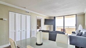 Un moderno salón con una mesa de comedor de cristal, un sofá gris y un televisor montado en la pared.