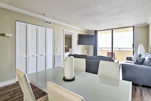A modern living room with a glass dining table, grey sofa, and a TV mounted on the wall. - Waterfront Condo: Resort Pool, Tennis & Beach (Hudson)