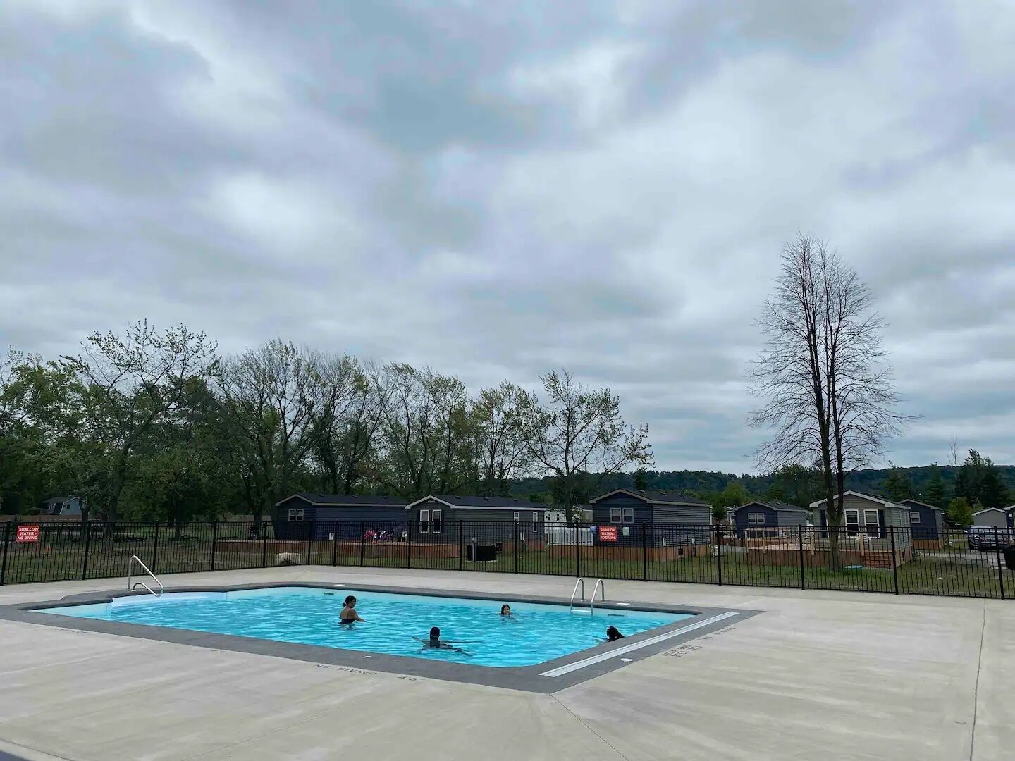 Piscina cercada por grade de metal, área pavimentada e árvores.