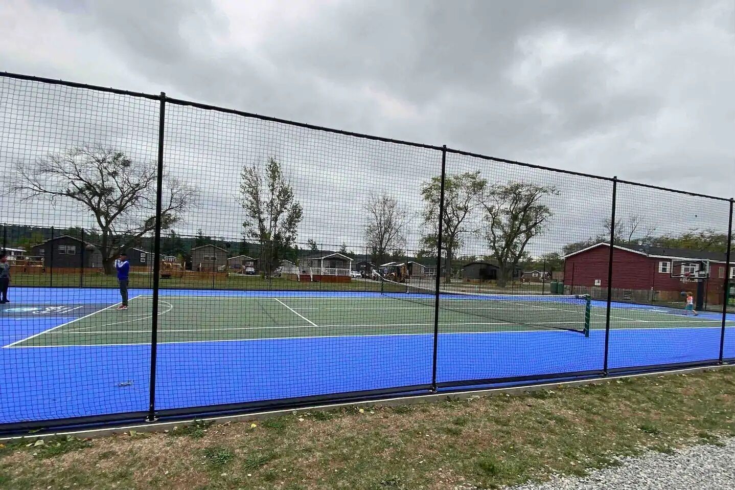 A tennis court with a net and surrounding fence, surrounded by houses and trees.