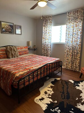 Une chambre à coucher comprenant un lit, un ventilateur de plafond, une fenêtre avec des rideaux, un tapis et des œuvres d’art murales.