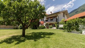 A two-story house with a red roof, surrounded by a well-maintained lawn and a large tree. - Large appartment with big garden, ideal for families! (Molini di Tures)
