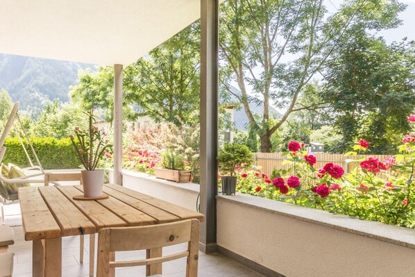 Mesa y sillas de madera bajo un porche cubierto, con vista a un jardín de flores rosas y árboles.