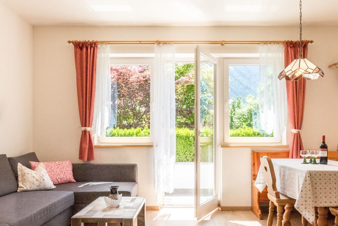 A cozy living room with a sofa, coffee table, and a dining area with a table and chairs. Large windows with curtains provide a view of greenery outside.