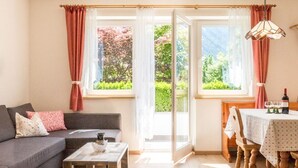 A cozy living room with a sofa, coffee table, and a dining area with a table and chairs. Large windows with curtains provide a view of greenery outside. - Large appartment with big garden, ideal for families! (Molini di Tures)