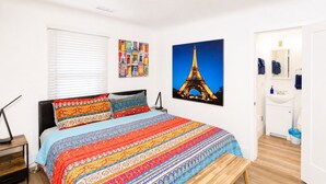 A bedroom with a bed, a bedside table, a nightstand, a window with blinds, a wall with a colorful artwork, and a bathroom visible through an open door. - Beaver Street Cottage w/AC, Fully Fenced Yard, W/D, 4 Smart TVs, Speedy Wi-Fi. (Flagstaff)