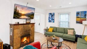 A living room with a stone fireplace, a green sofa, a red chair, and a flat-screen TV mounted above the fireplace. - Beaver Street Cottage w/AC, Fully Fenced Yard, W/D, 4 Smart TVs, Speedy Wi-Fi. (Flagstaff)