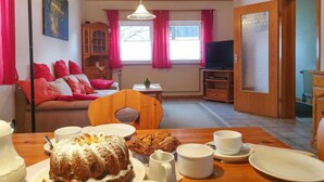 A cozy living room with a wooden ceiling, a sofa with red cushions, a dining table set with plates, cups, and a cake.