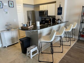 Una cocina moderna con una isla central, taburetes blancos tipo bar, un refrigerador de acero inoxidable y un microondas.