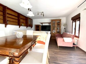 A spacious living area with a wooden dining table, a pink sofa, and a television mounted on the wall.