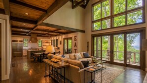 A spacious living room with a large window overlooking a forested area, a kitchen with modern appliances, and a cozy seating area with a fireplace. - Timber Forge Lodge - Eagles Nest home with views, hot tub, game table, more! (Banner Elk)