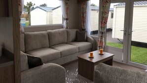 A cozy living area with a sofa, a small table with candles, and a view of a residential neighborhood through large windows. - 3 Bed Luxury Mobile Home Hoburne Park Christchurch (Christchurch)