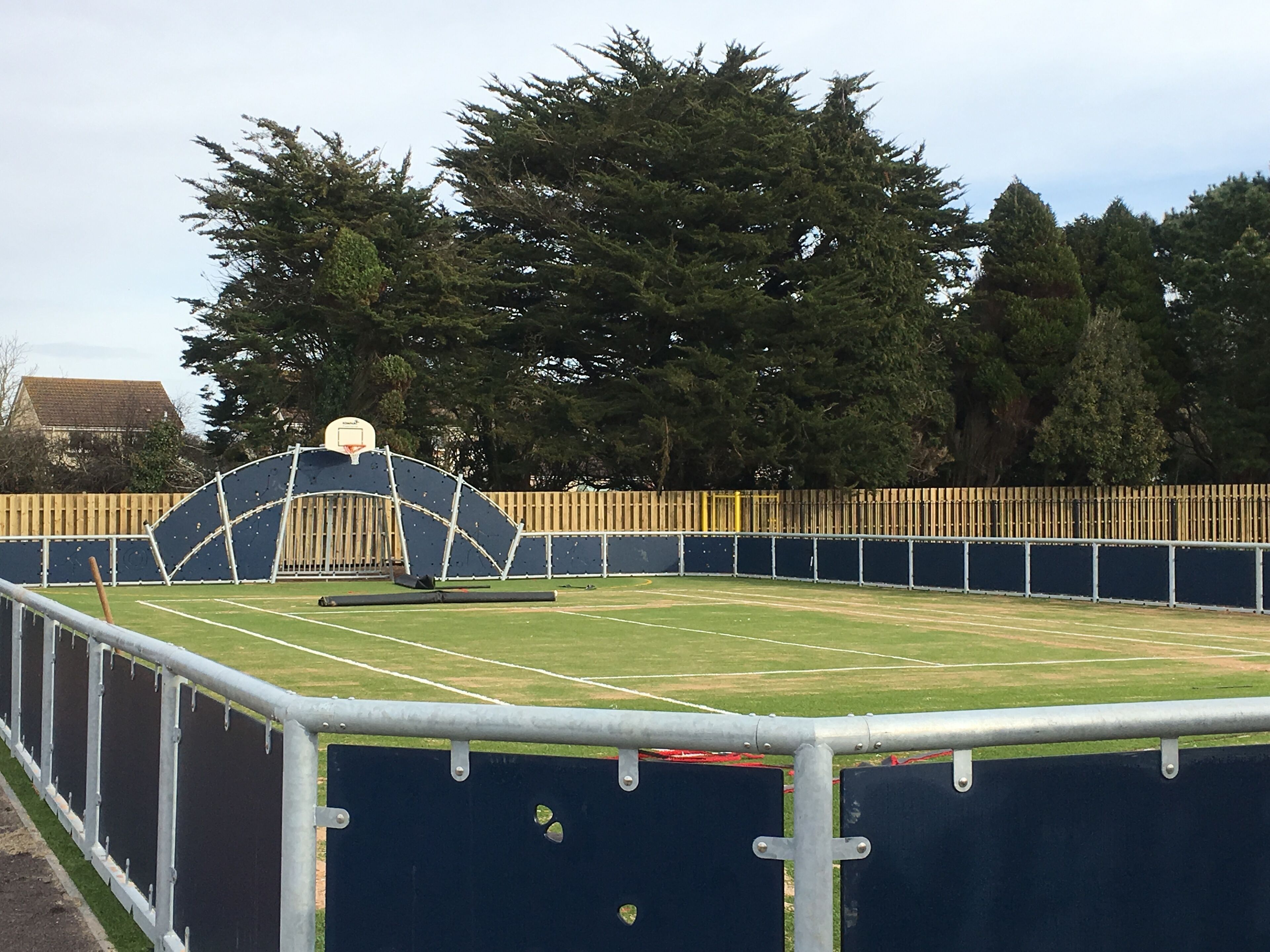 A tennis court with a metal fence, trees, and a basketball hoop in the background.