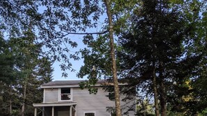 A two-story cabin with a porch, surrounded by trees and a grassy area. - Jameson Point Cottage in Scenic Friendship, Me (Friendship)