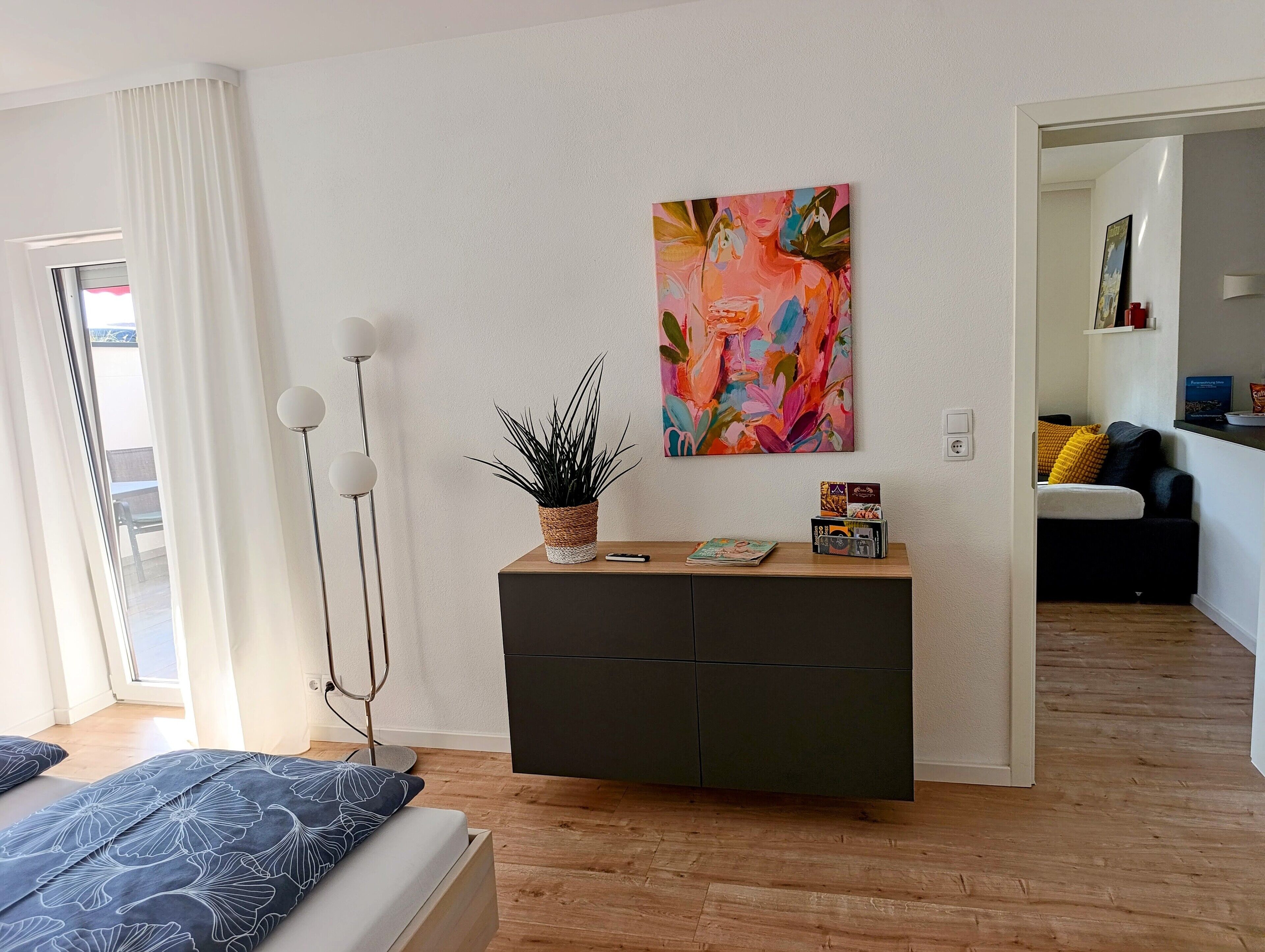 Ein modernes Wohnzimmer mit einem Bett, einem Konsolentisch mit Pflanze, einem farbenfrohen abstrakten Gemälde und Blick auf einen Balkon.